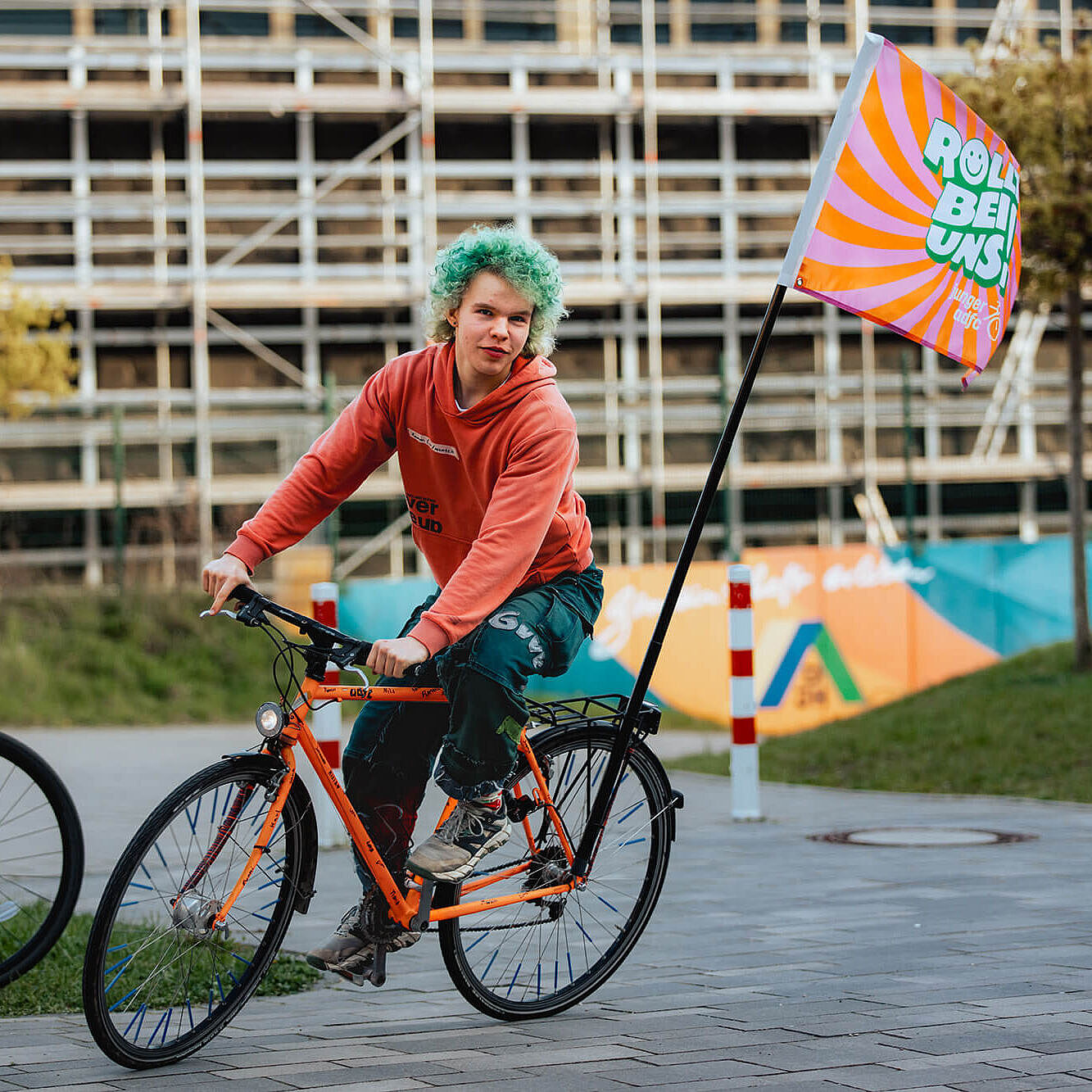 Eine junge Person fährt auf einem orangefarbenen Fahrrad. Hinten am Rad weht eine bunte Fahne des Jungen ADFC.