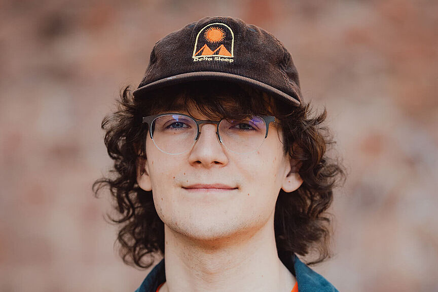 Elija König Porträt eines jungen Mannes mit Brille und dunkler Kappe. Er trägt ein orangefarbenes Shirt des Jungen ADFC und schaut in die Kamera.