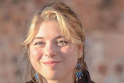 Porträt einer jungen Frau im Sonnenlicht. Sie sitzt draußen und schaut mit einem leichten Lächeln in die Kamera.