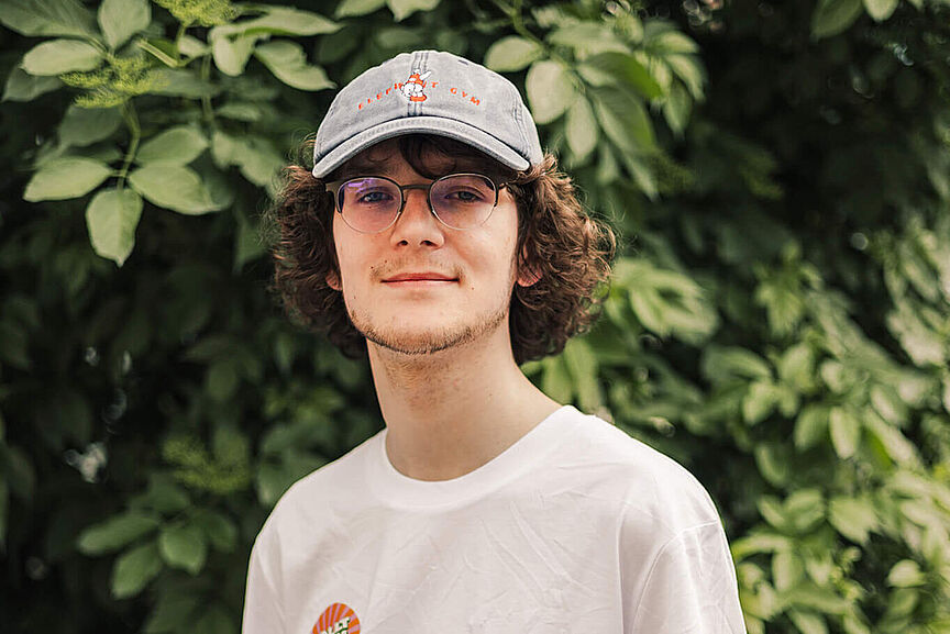 Elija König Junger lächelnder Mann mit weißem T-Shirt mit Aufdruck Rollt bei uns, Brille und einem jeansblauen Cappy vor grünem Blätterhintergrund.