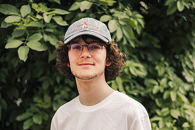 Junger lächelnder Mann mit weißem  T-Shirt mit Aufdruck Rollt bei uns, Brille und einem jeansblauen Cappy vor grünem Blätterhintergrund.