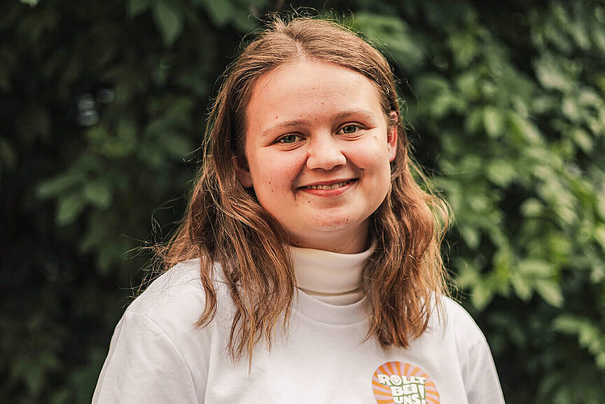 Lilia Vogt Lächelnde Frau mit weißem T-Shirt und Aufdruck ‚Rollt bei uns!‘ vor grünem Hintergrund.