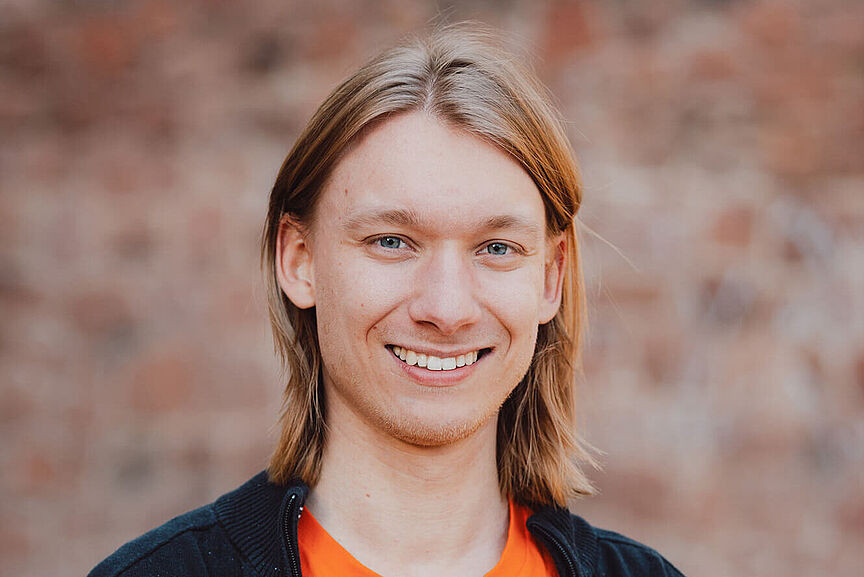 Sebastian Vogel Porträt eines jungen Mannes im Freien. Er trägt ein orangefarbenes Shirt mit dem Jungen ADFC Logo und eine dunkle Jacke. Er lächelt in die Kamera.