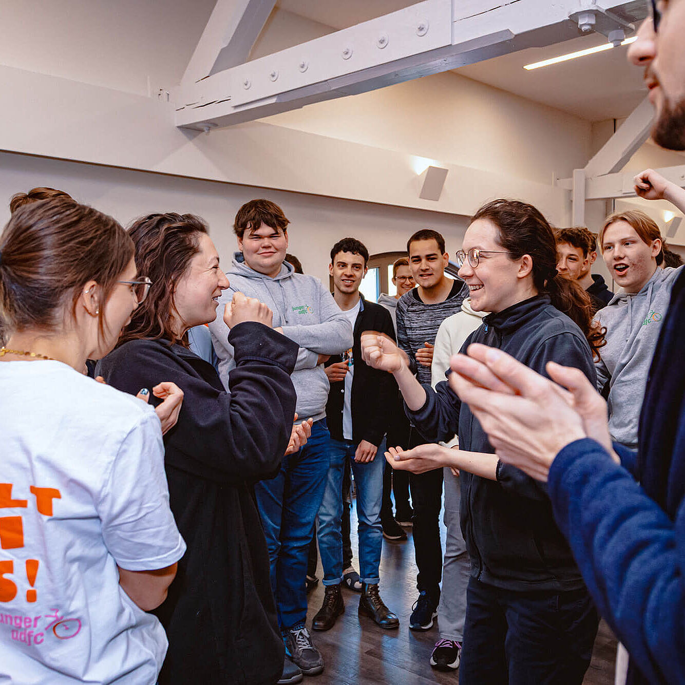 Mehrere junge Menschen stehen in einem Raum zusammen und lachen. Zwei Personen spielen in der Mitte miteinander, die anderen schauen zu und klatschen.