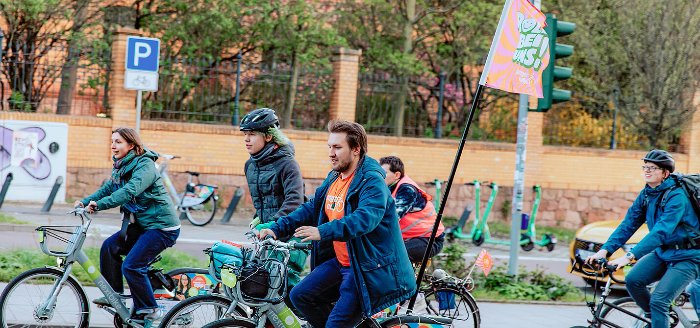NRVP Halbzeitbilanz Mehrere Radfahrende sind auf einer Straße unterwegs. Eine Person hält eine Fahne, im Hintergrund stehen Bäume und Verkehrsschilder.