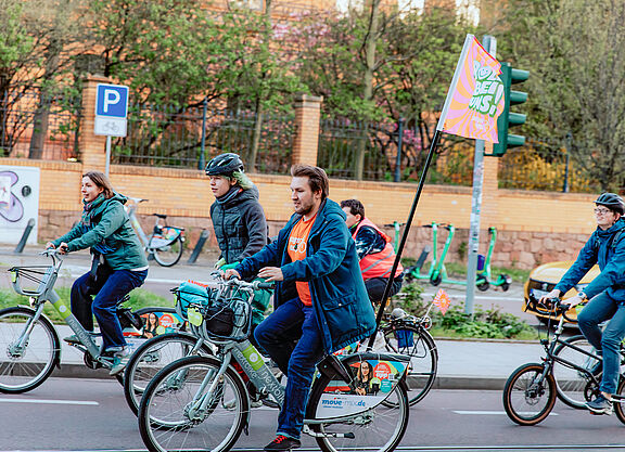 Mehrere Radfahrende sind auf einer Straße unterwegs. Eine Person hält eine Fahne, im Hintergrund stehen Bäume und Verkehrsschilder.