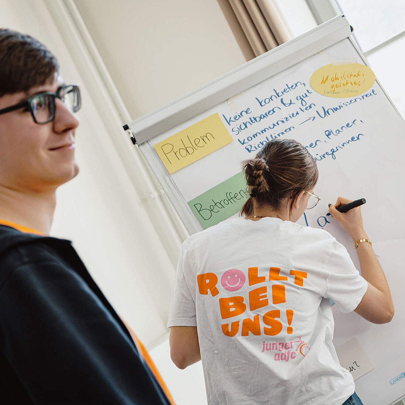Eine junge Person schreibt an ein großes Flipchart. Daneben steht ein weiterer junger Mensch und schaut lächelnd zu.