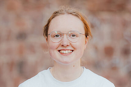 Porträt einer jungen Frau mit Brille. Sie lächelt in die Kamera. Im Hintergrund ist eine unscharfe Wand aus Ziegeln zu sehen.