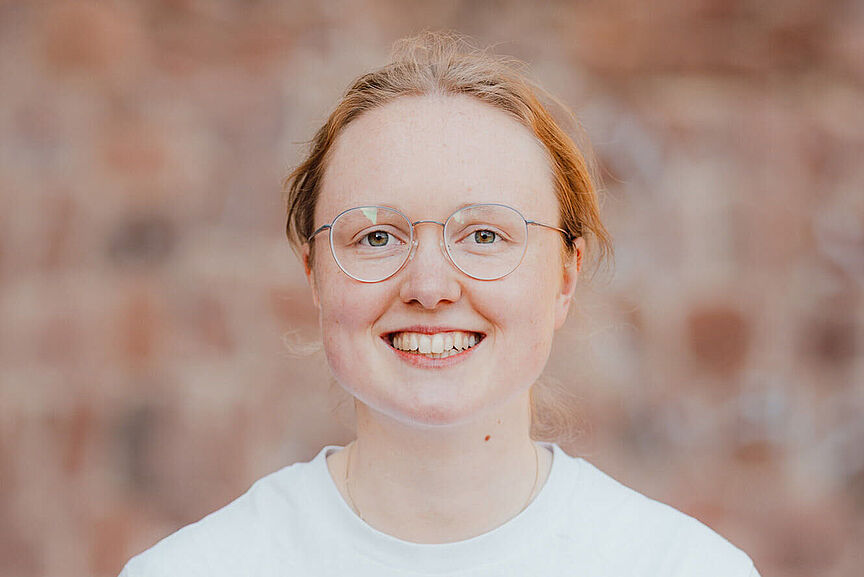 Lena Becker Porträt einer jungen Frau mit Brille. Sie lächelt in die Kamera. Im Hintergrund ist eine unscharfe Wand aus Ziegeln zu sehen.