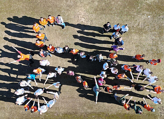 Gruppe von Menschen steht auf einem Platz und bildet gemeinsam die Form eines Fahrrads. Die Personen tragen teils orangefarbene, teils weiße oder bunte Oberteile. Das Bild wurde aus der Vogelperspektive aufgenommen.