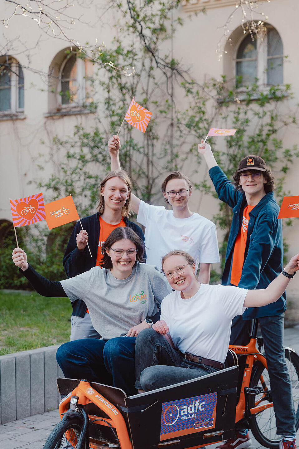 Bundesjugendvorstand Fünf junge Menschen posieren draußen auf und neben einem Lastenrad. Sie lachen und halten kleine Fahnen des Jungen ADFC in die Höhe.