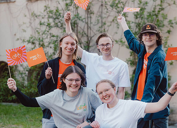 Fünf junge Menschen posieren draußen auf und neben einem Lastenrad. Sie lachen und halten kleine Fahnen des Jungen ADFC in die Höhe.