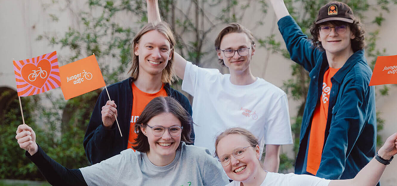 Fünf junge Menschen posieren draußen auf und neben einem Lastenrad. Sie lachen und halten kleine Fahnen des Jungen ADFC in die Höhe.