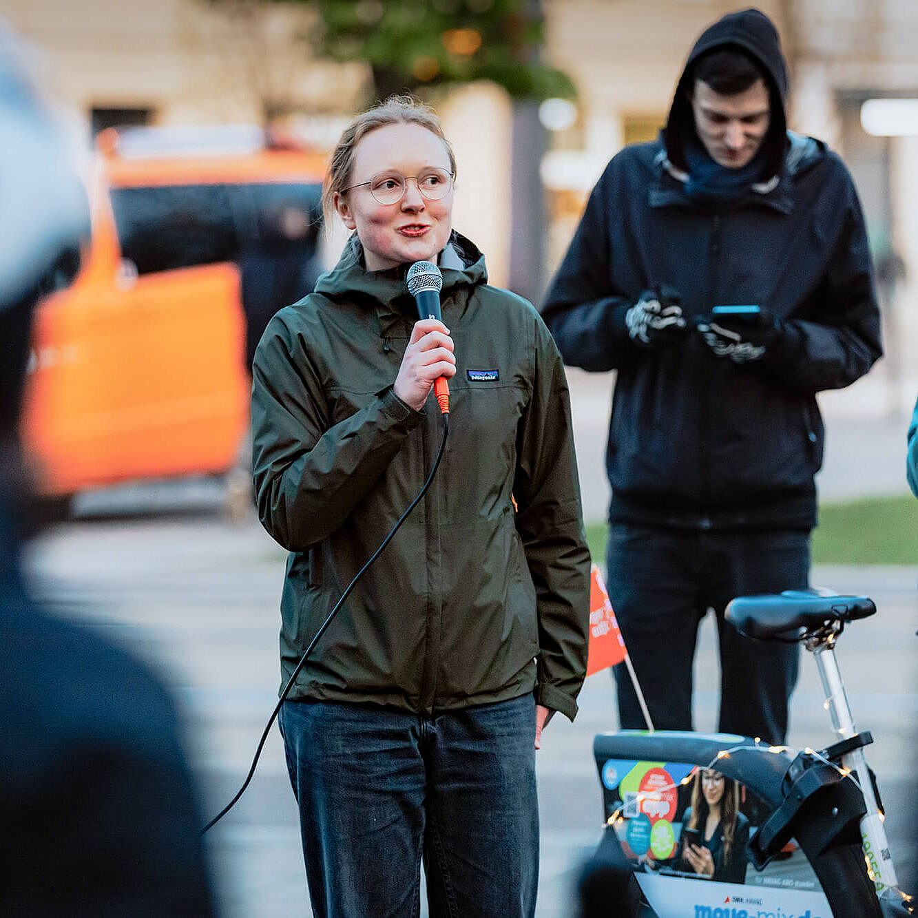 Eine junge Frau steht draußen mit einem Mikrofon in der Hand und spricht zu einer Gruppe. Neben ihr steht ein Fahrrad, im Hintergrund schauen weitere Menschen zu.