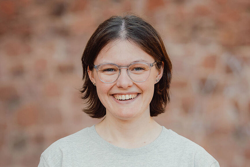 Lena Adam Porträt einer jungen Frau mit Brille. Sie trägt ein graues Shirt des Jungen ADFC und lächelt in die Kamera.