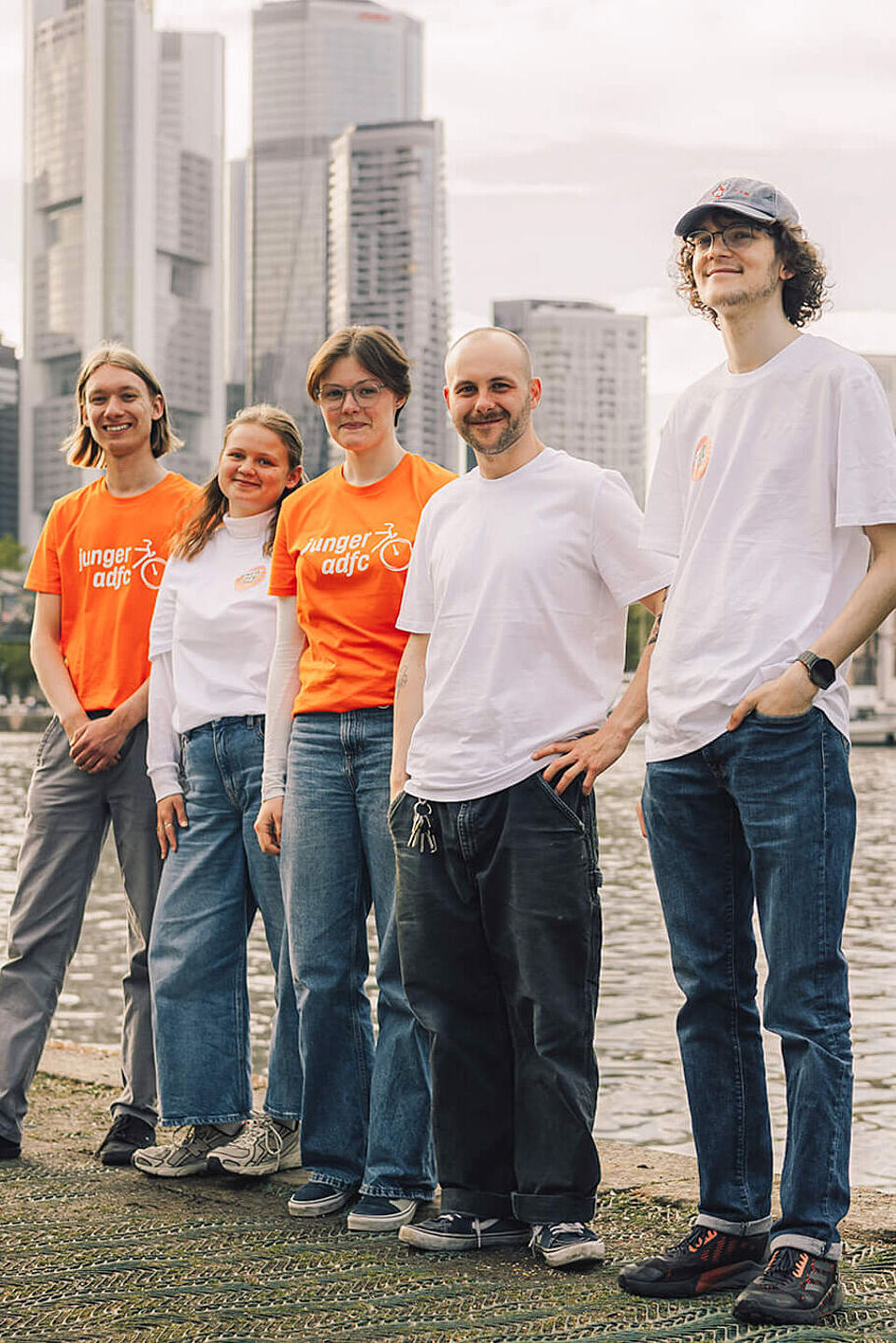 Bundesjugendvorstand - Junger ADFC Fünf junge Menschen stehen am Mainufer in Frankfurt, drei mit orangen ‚Junger ADFC‘-T-Shirts, zwei in weißen T-Shirts, im Hintergrund Skyline und Eiserner Steg.