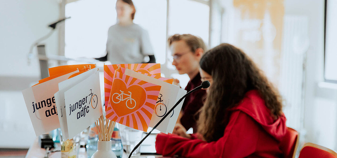 Bundesjugendversammlung 2026 Auf einem Tisch stehen kleine Fahnen mit dem Logo des jungen ADFC. Daneben sitzen junge Menschen an Mikrofonen in einem hellen Raum.