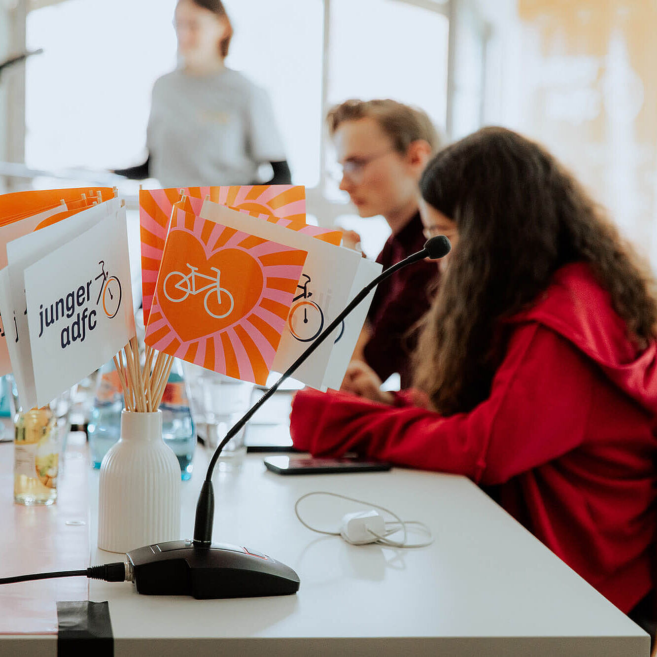 Auf einem Tisch stehen kleine Fahnen mit dem Logo des jungen ADFC. Daneben sitzen junge Menschen an Mikrofonen in einem hellen Raum.