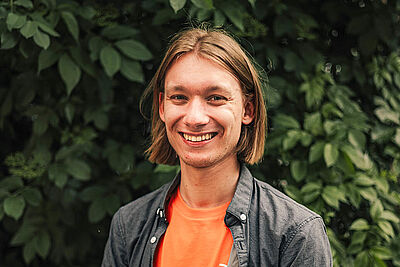 Junger Mann mit schulterlangen Haaren, orangenem „Junger ADFC“-T-Shirt und offener, dunkelgrauer Hemdjacke steht lächelnd vor einer dichten, grünen Blattkulisse.