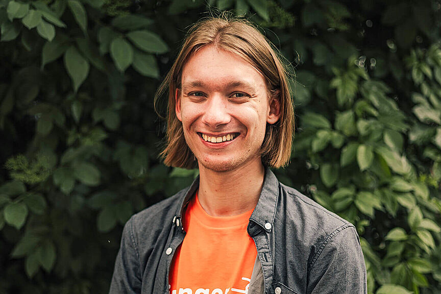 Sebastian Vogel Junger Mann mit schulterlangen Haaren, orangenem „Junger ADFC“-T-Shirt und offener, dunkelgrauer Hemdjacke steht lächelnd vor einer dichten, grünen Blattkulisse.