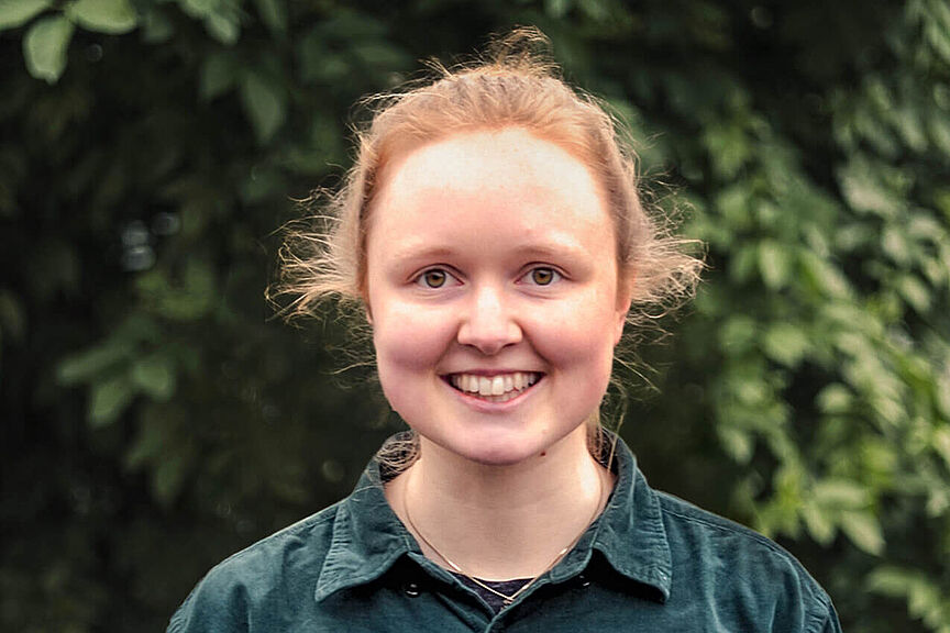 Lena Becker Lächelnde Frau im dunkelgrünen Hemd vor grünem Blätterhintergrund.