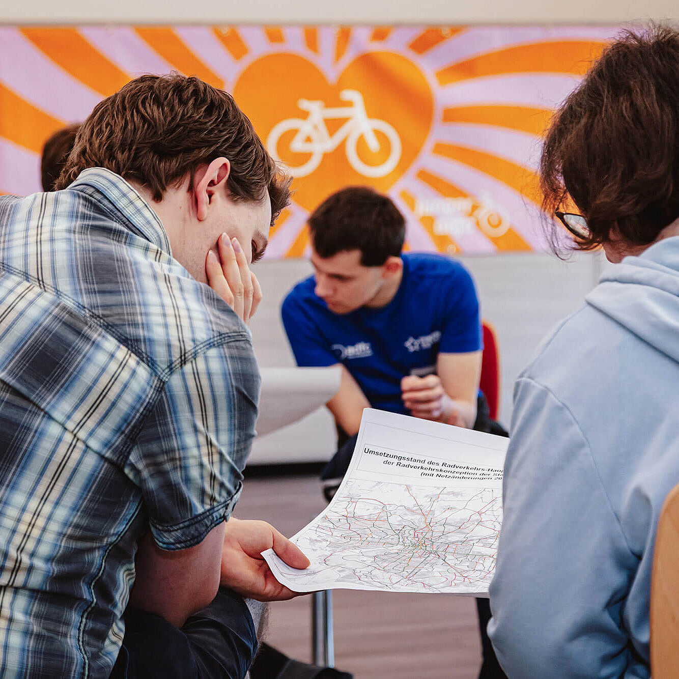 Zwei junge Menschen sitzen in einem Raum und schauen auf einen Plan. Im Hintergrund spricht eine weitere Person vor einer Wand mit dem Logo des Jungen ADFC.