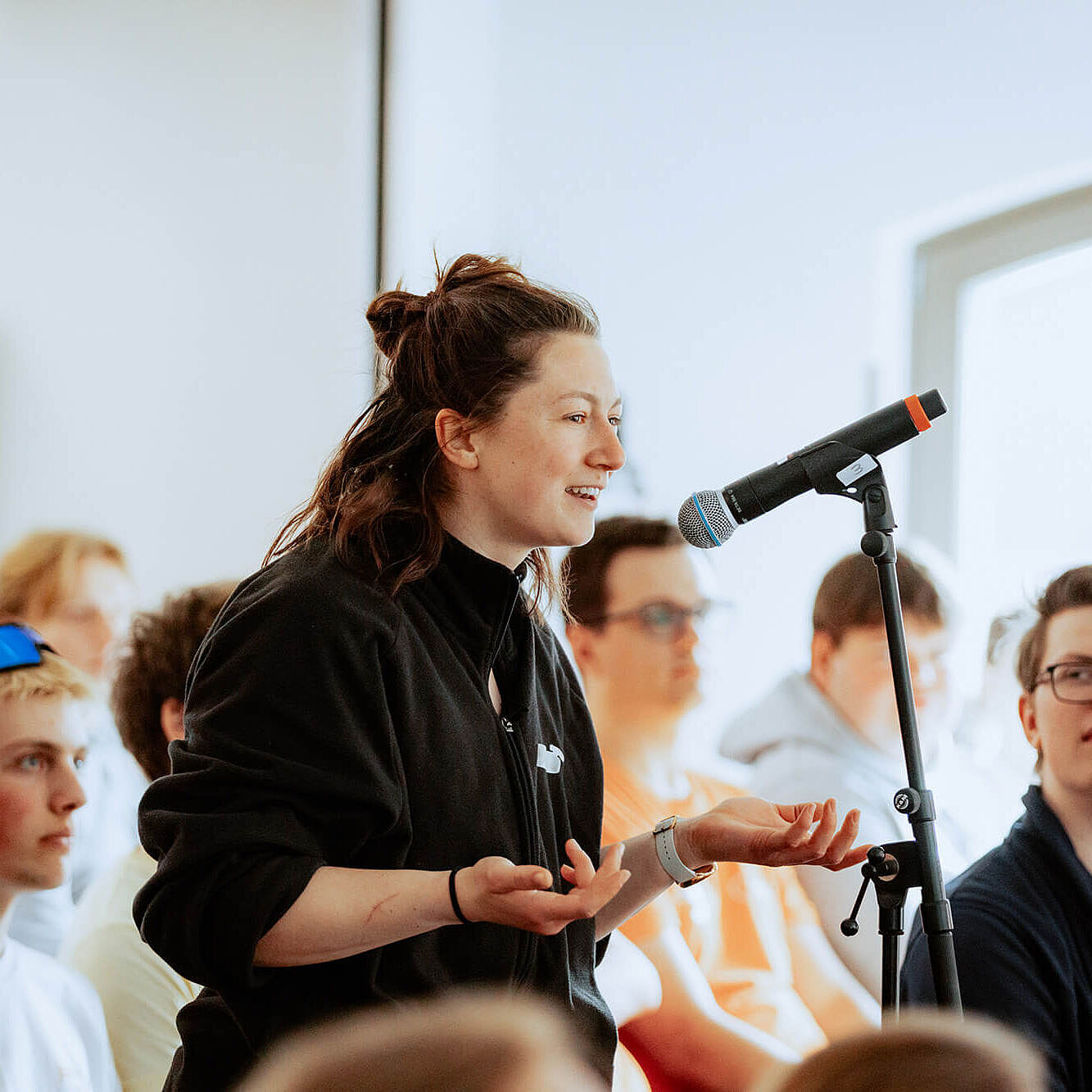 Eine junge Frau spricht in ein Mikrofon in einem hellen Raum. Um sie herum sitzen viele junge Menschen und hören zu.