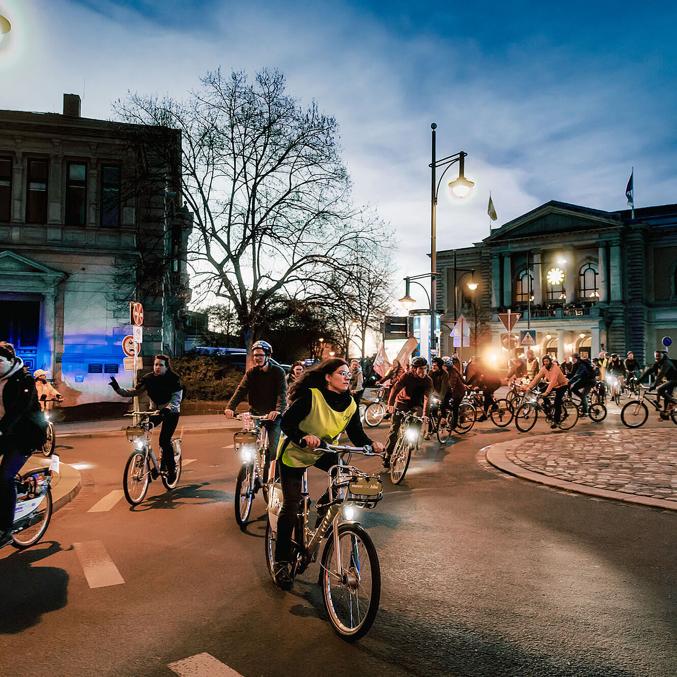 Viele Menschen fahren am Abend mit Fahrrädern durch eine Stadt. Straßenlaternen und Fahrradlichter leuchten, im Hintergrund stehen große Gebäude.
