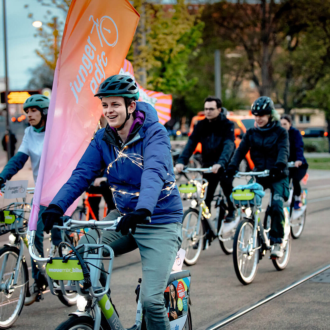 Mehrere Menschen fahren am Abend mit Fahrrädern durch die Stadt. Vorne fährt ein junger Mann mit Helm neben einer großen Fahne des Jungen ADFC.