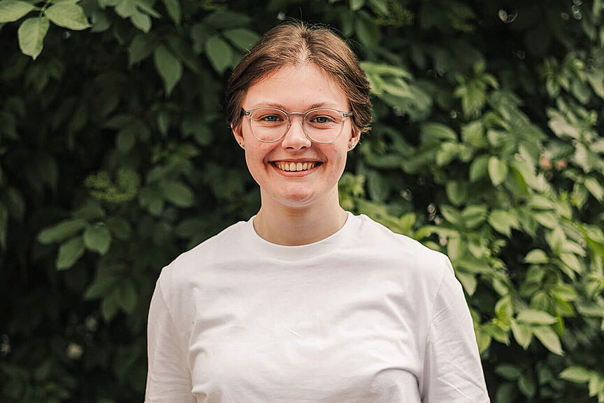 Lena Adam Junge lächelnde Frau mit weißem Langarmshirt unter weißem T-Shirt und Brille vor grünem Blätterhintergrund.