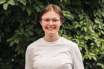 Junge lächelnde Frau mit weißem Langarmshirt unter weißem T-Shirt und Brille vor grünem Blätterhintergrund.