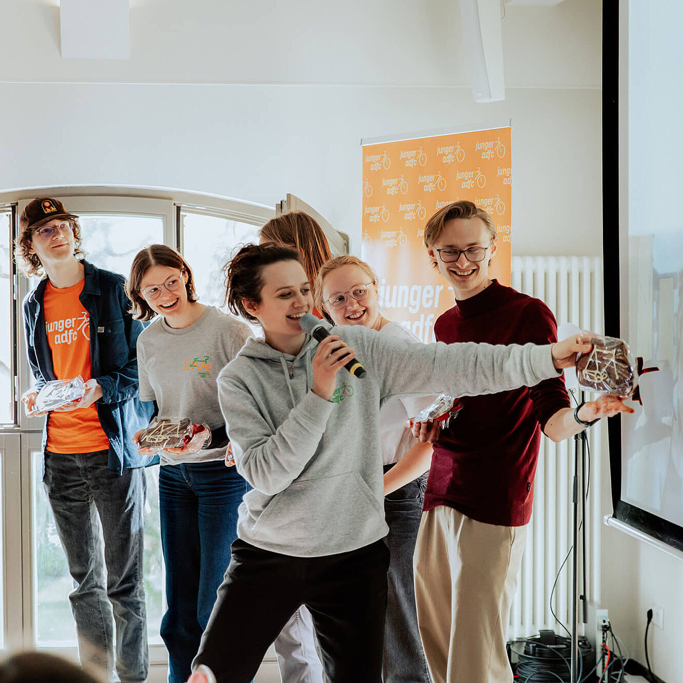 Mehrere junge Menschen stehen vor einer Leinwand. Eine Person spricht ins Mikrofon und zeigt auf ein Bild, die anderen lachen.