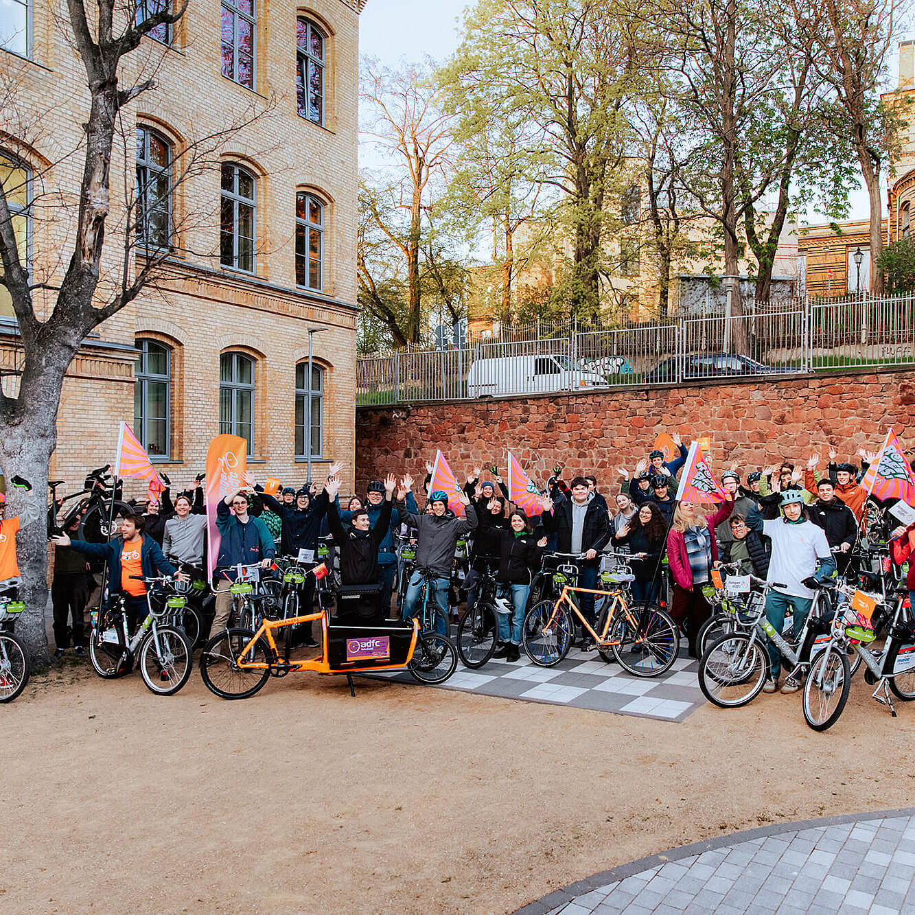 Viele junge Menschen stehen mit Fahrrädern vor einem großen Gebäude im Freien. Einige halten Fahnen hoch und winken in die Kamera.