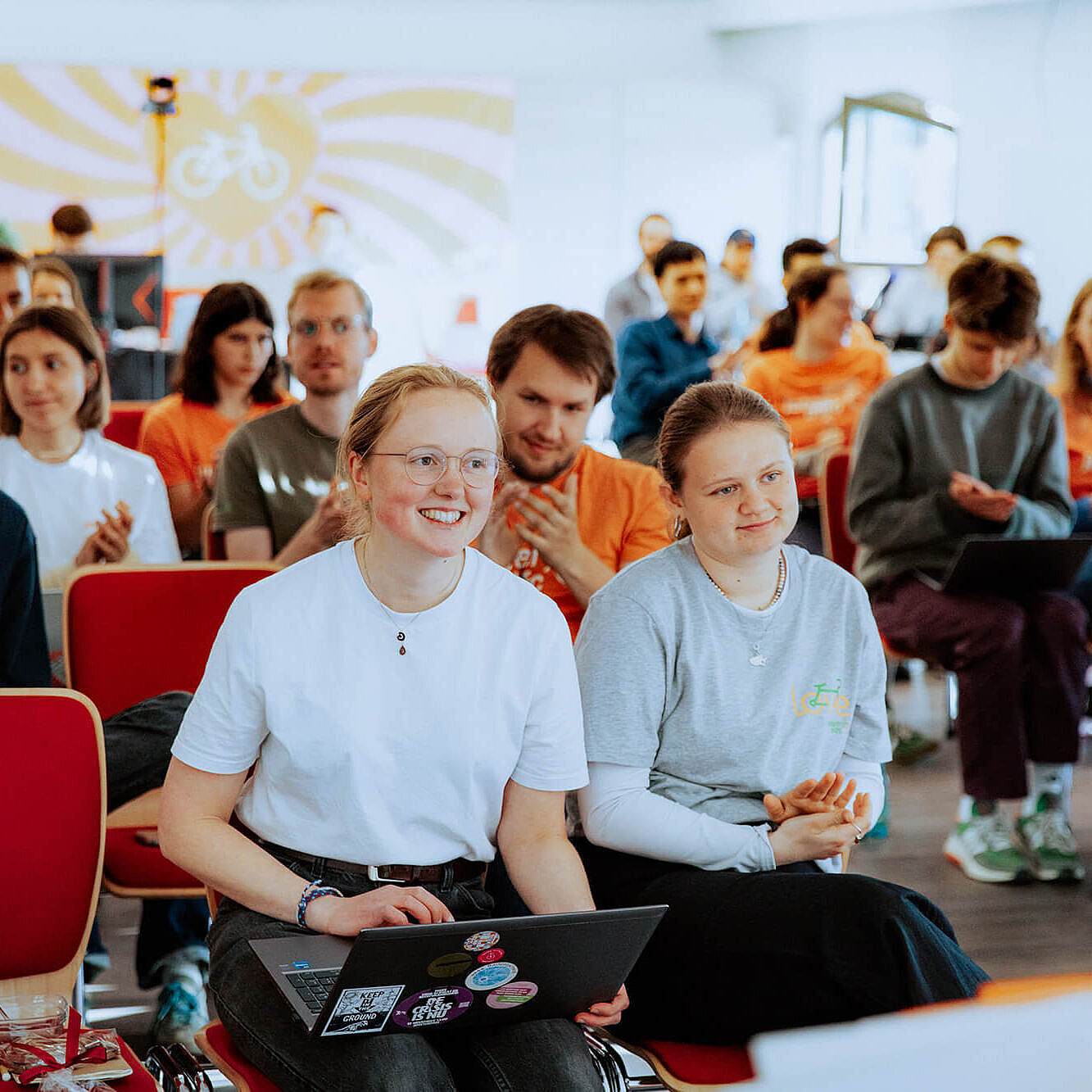 Viele junge Menschen sitzen in einem hellen Raum auf roten Stühlen. Vorne sitzen zwei Personen mit einem Laptop auf dem Schoß.